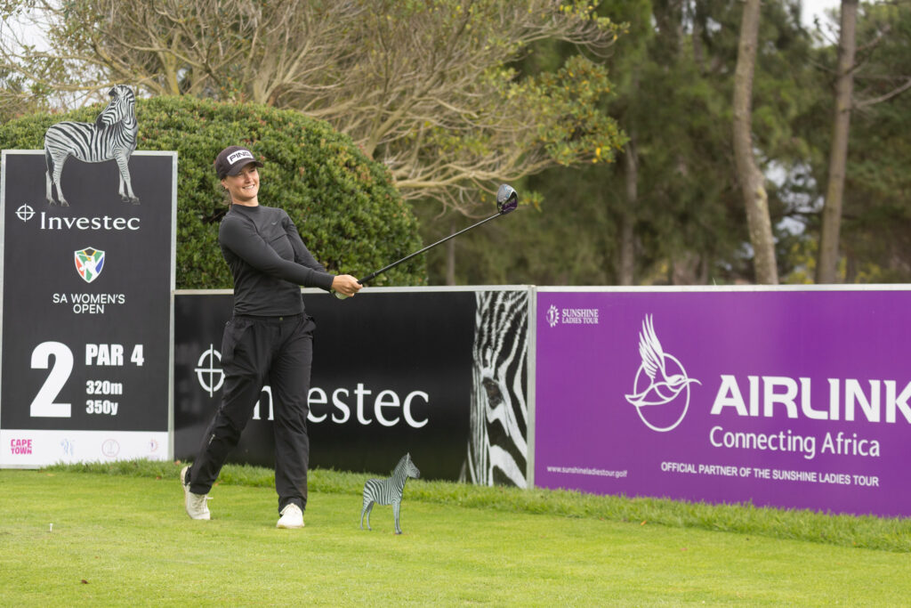 Female golfer in black swings a club on a grassy course with zebra-themed sponsor boards in the background.