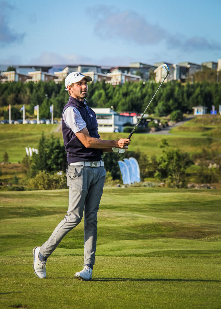 Male golfer in a navy vest and white cap swings a club on a sunny golf course with green fairways and buildings in the background.