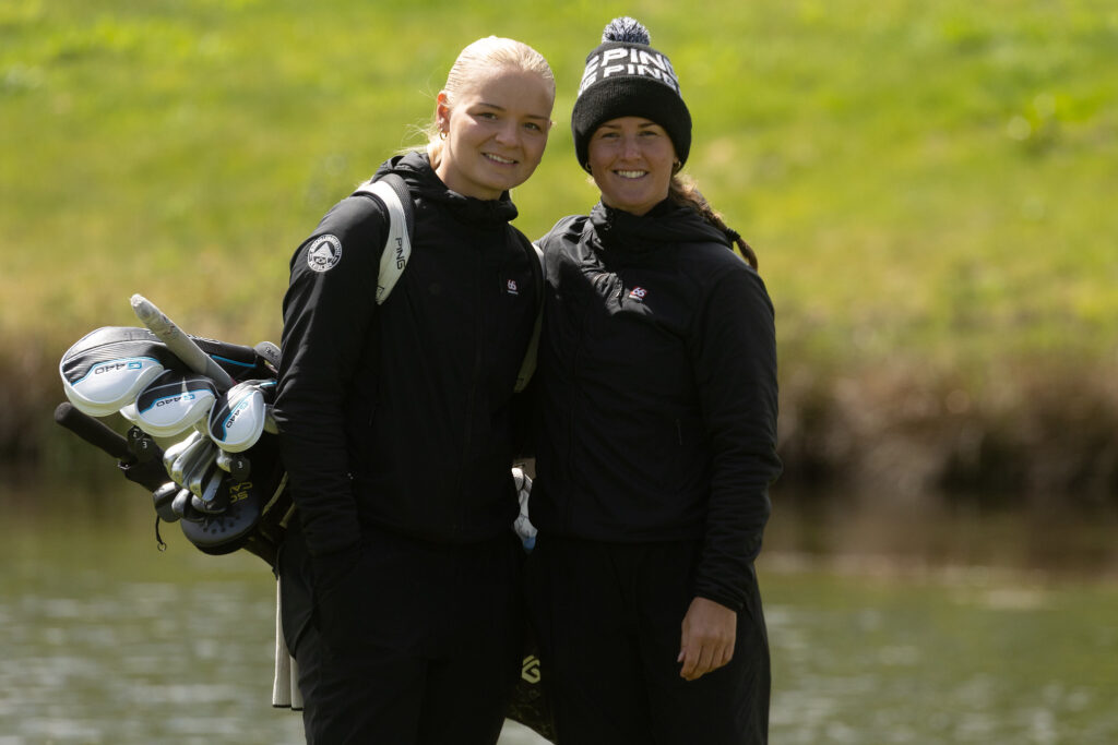 Two women in black jackets stand together outdoors, one carrying a golf bag full of clubs by a pond or lake.