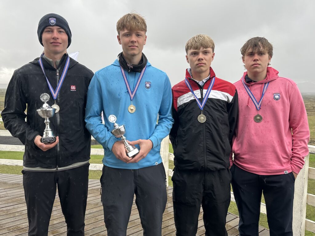 Four young men standing side by side on a wooden deck, each wearing medals; two hold trophies after a sports event, cloudy sky above.