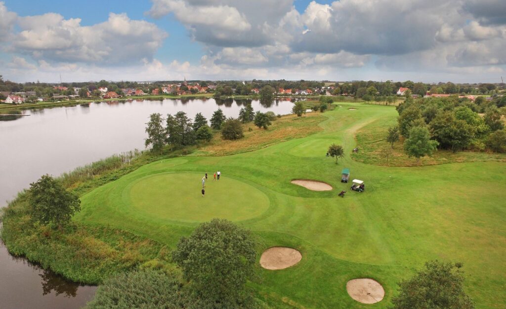 Aerial view of a golf course by a lake with players on the green and sand bunkers nearby, houses in the distance.