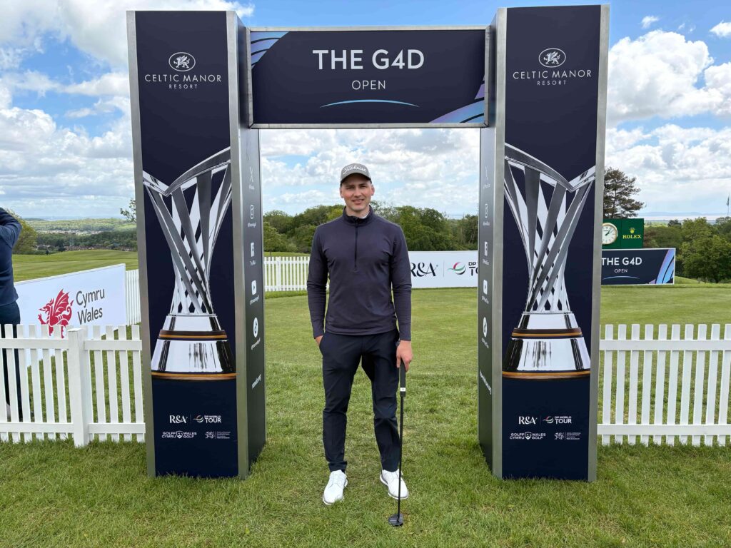 Man in dark sportswear standing with a golf club under an archway that says THE G4D OPEN, Celtic Manor banners visible in a field event setting