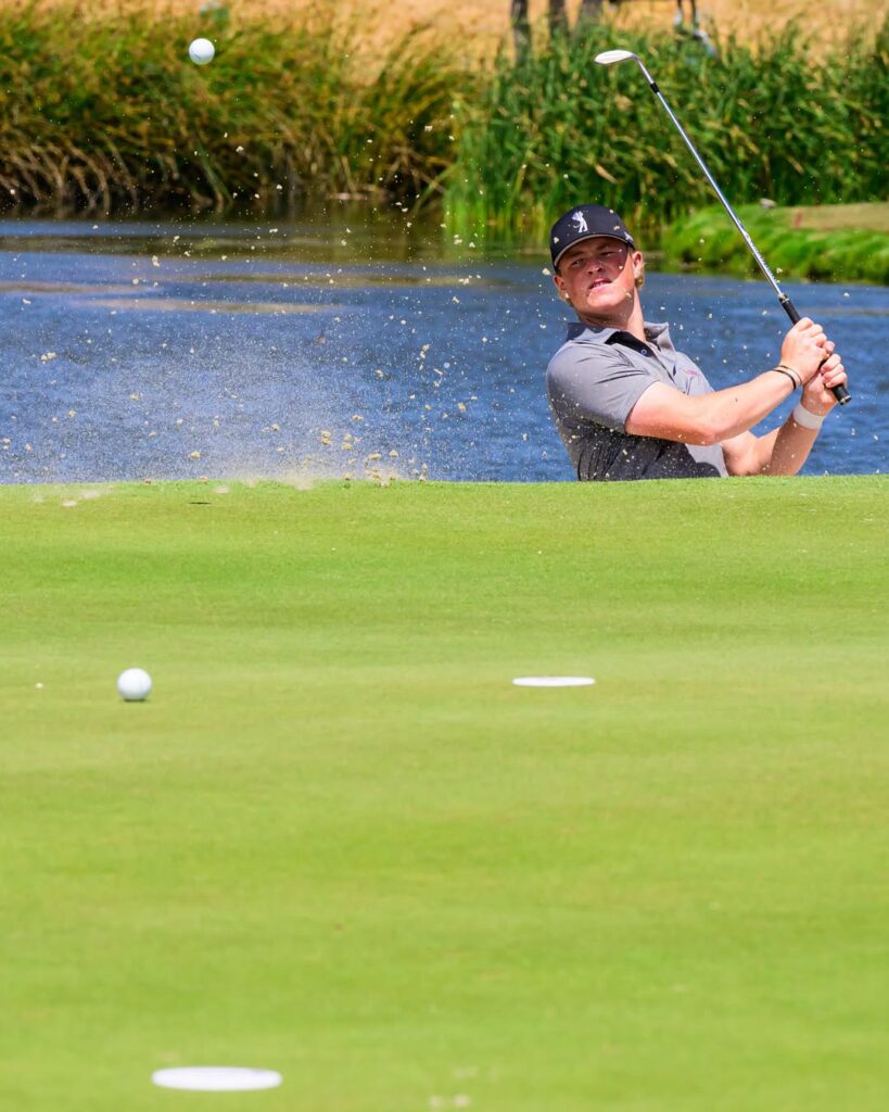 Male golfer in a gray polo and cap hits a shot from a sand bunker with water behind, ball rising through the air.