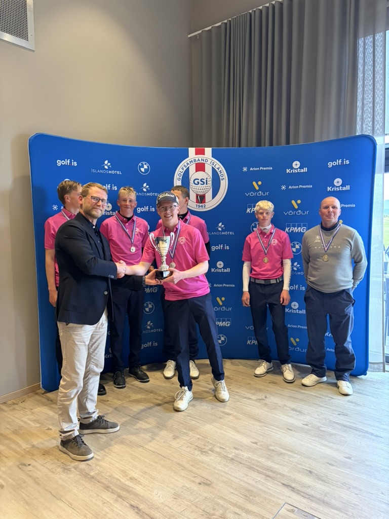 Team of young golfers in pink shirts pose with a trophy and medals in front of a blue sponsor backdrop.
