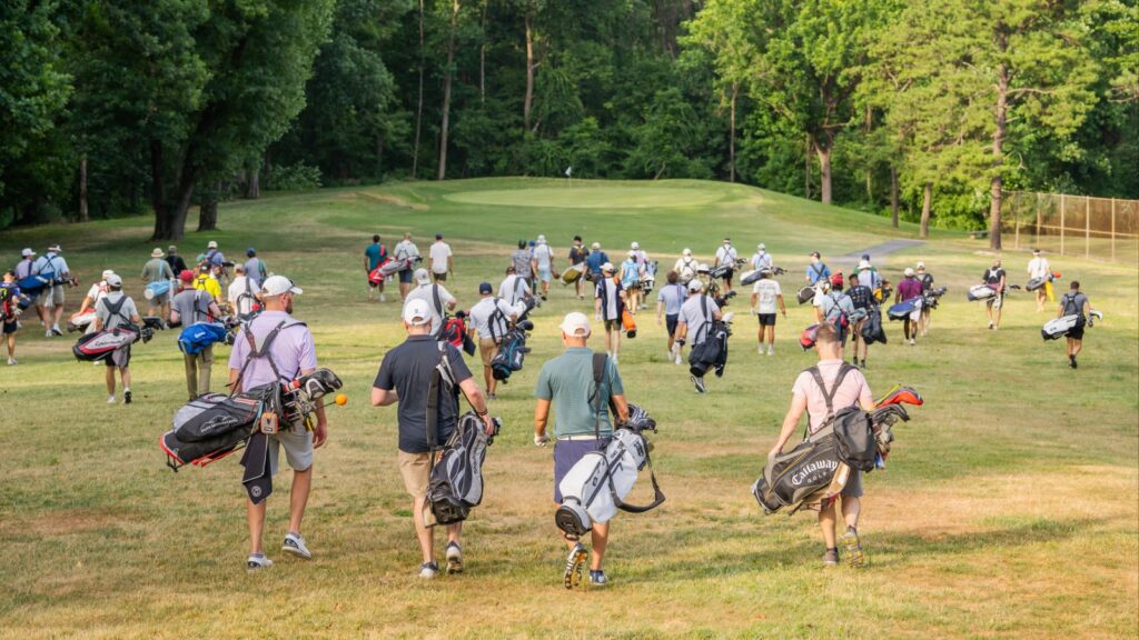 Group of golfers walking across a fairway toward the green, each carrying a golf bag on their backs or shoulders.
