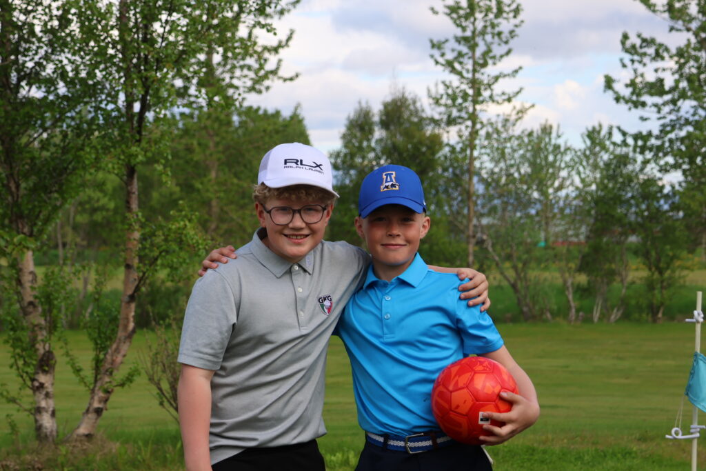 Two boys outdoors with trees in the background, arms around each other, one holding a red soccer ball central to the frame.