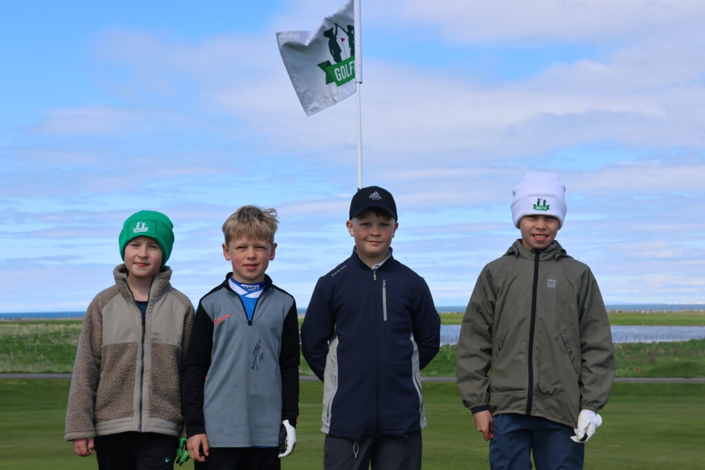Five young golfers stand side by side on a grassy golf course, flagpole behind them against a blue cloudy sky.