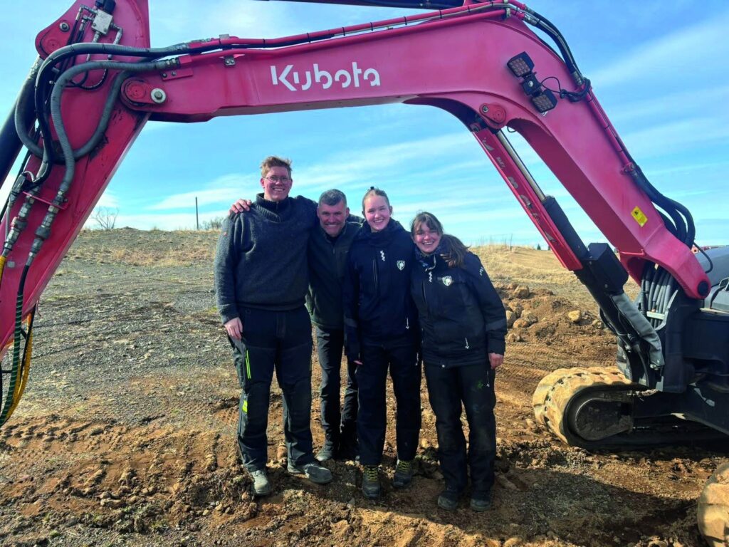 Four workers in workwear pose together in front of a pink Kubota excavator on a muddy construction site, smiling at the camera.