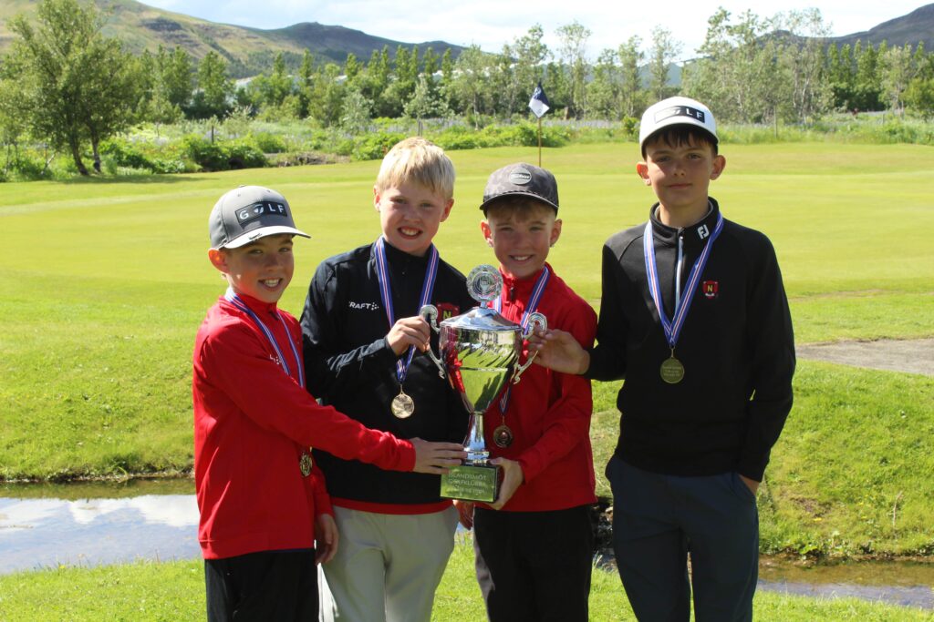 Four boys in red and black sportswear pose with medals and a large trophy on a golf course.