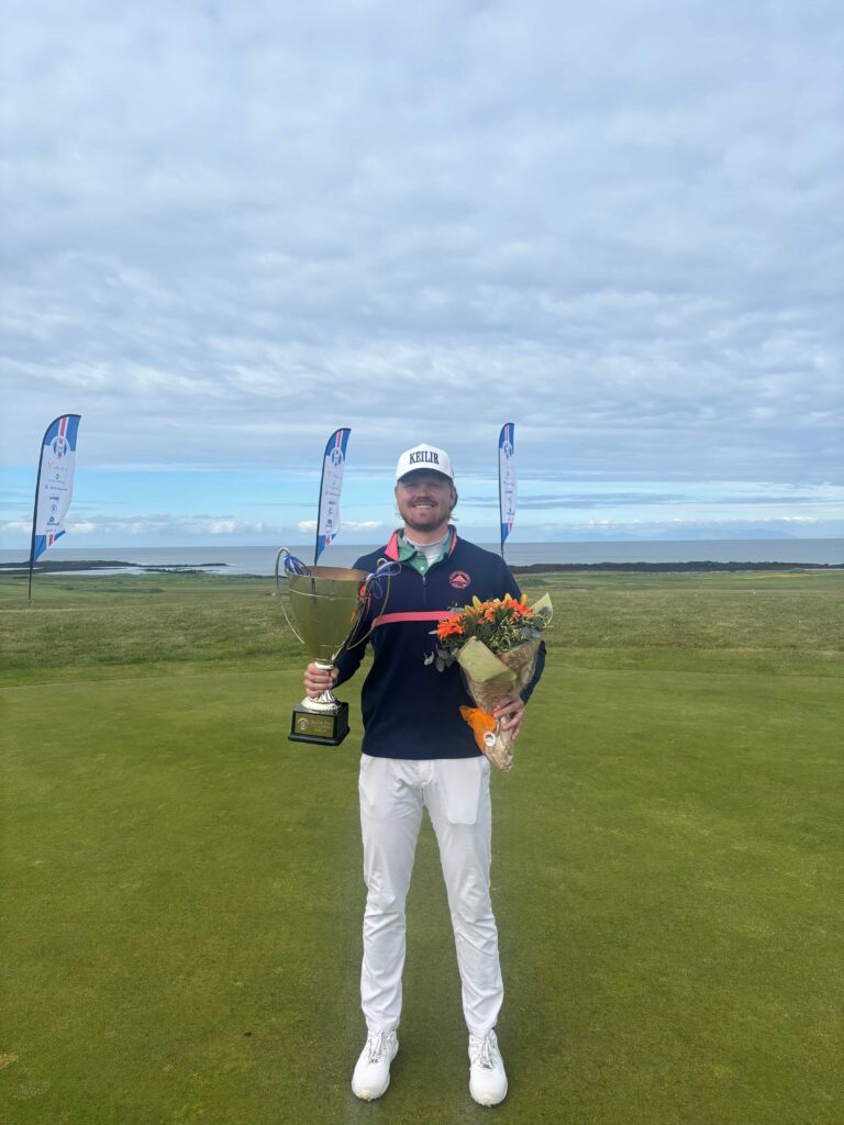 Golfer standing on a course holding a large trophy in one hand and a bouquet of flowers in the other, with banners in the background and a coastal landscape behind him.