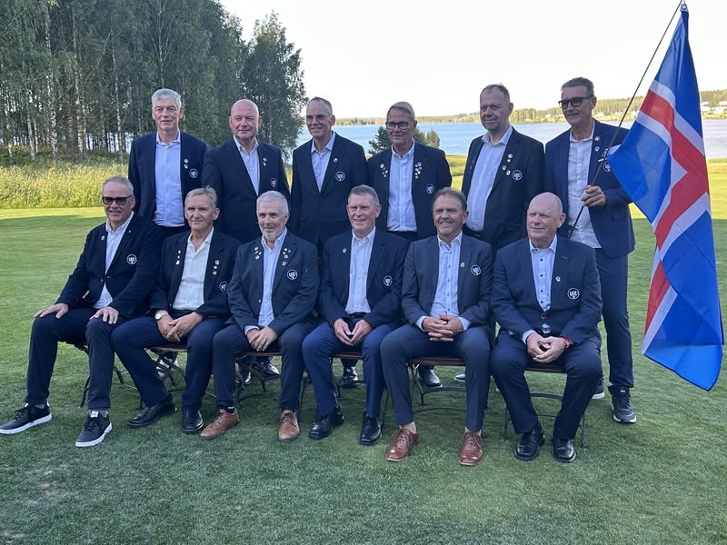 Group of men in matching blazers posing for a formal photo on a golf course beside a blue, white, and red flag.
