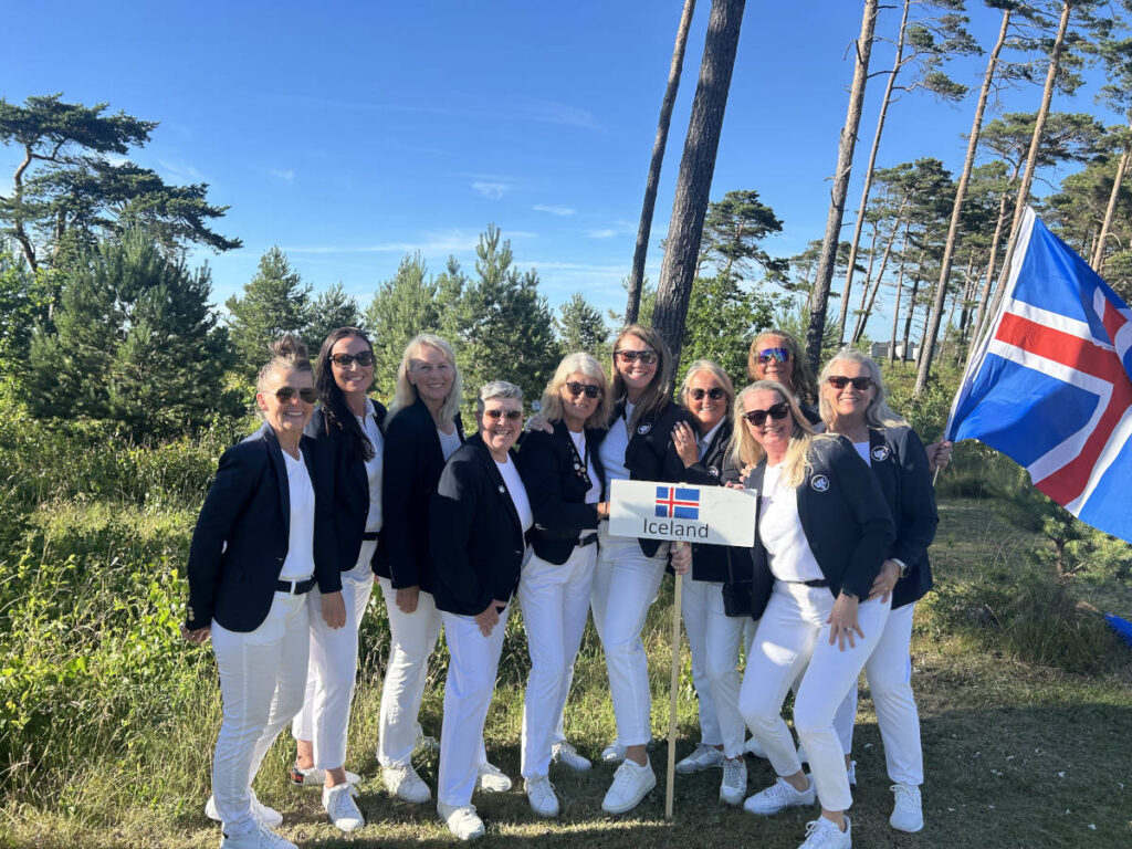 Group of ten women in white pants and navy blazers posing outdoors with an Iceland sign and flag in a pine forest under a blue sky.