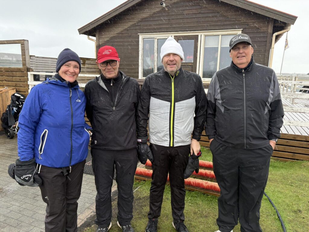 Four adults in waterproof jackets and beanies stand side by side on a wet boardwalk in front of a brown wooden cabin, posing for a photo in the rain with red cleaning brushes on the ground nearby.