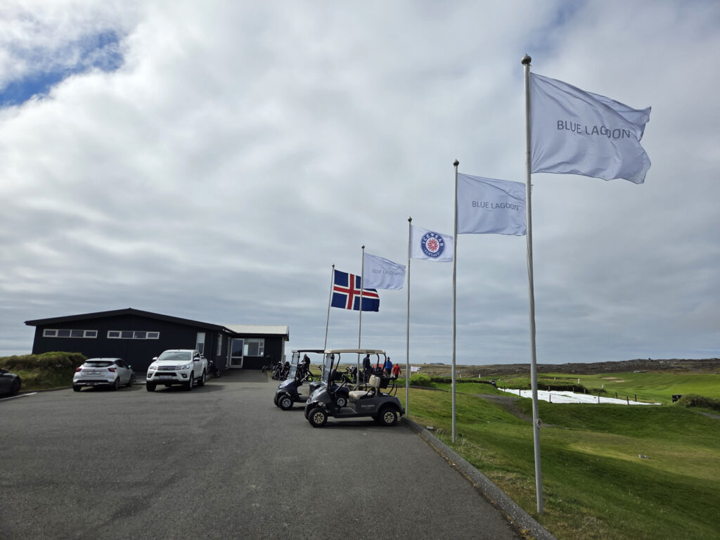 Flagpoles with Blue Lagoon banners and the Icelandic flag beside a dark modern building, with parked cars and golf carts on a paved driveway at a coastal resort.