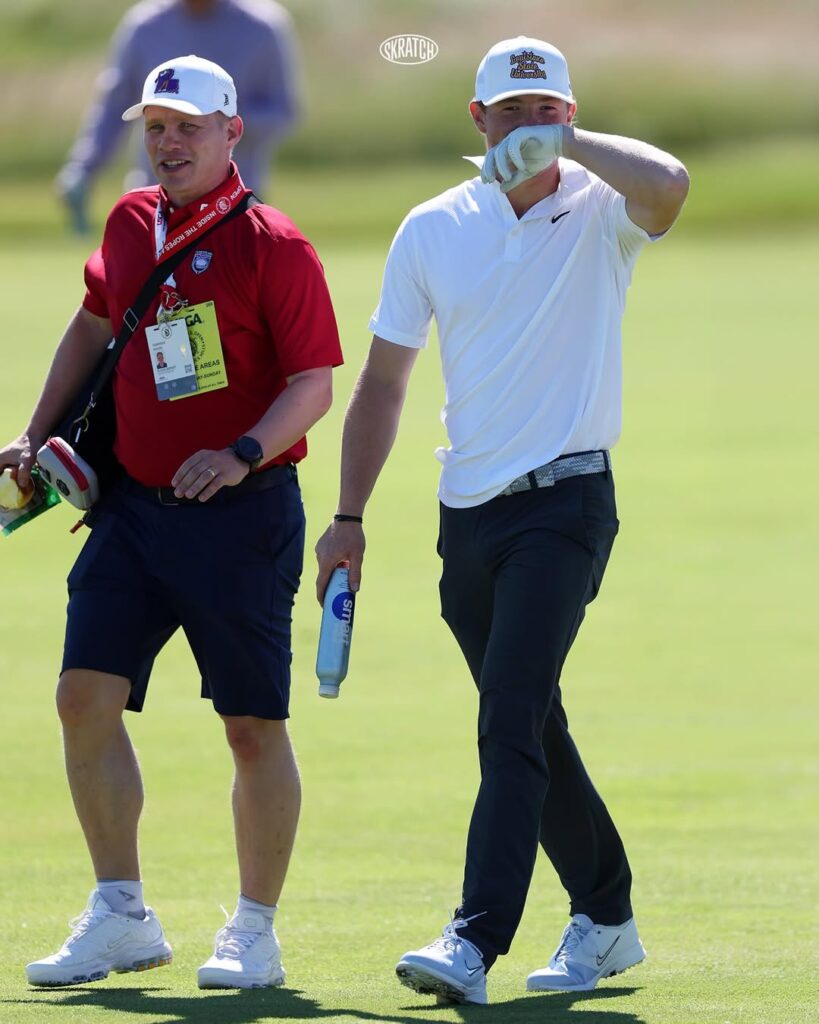 Two male golfers walk on a sunny green: one in a red polo with badges and gear, the other in a white polo shielding his eyes with a gloved hand and carrying a water bottle.