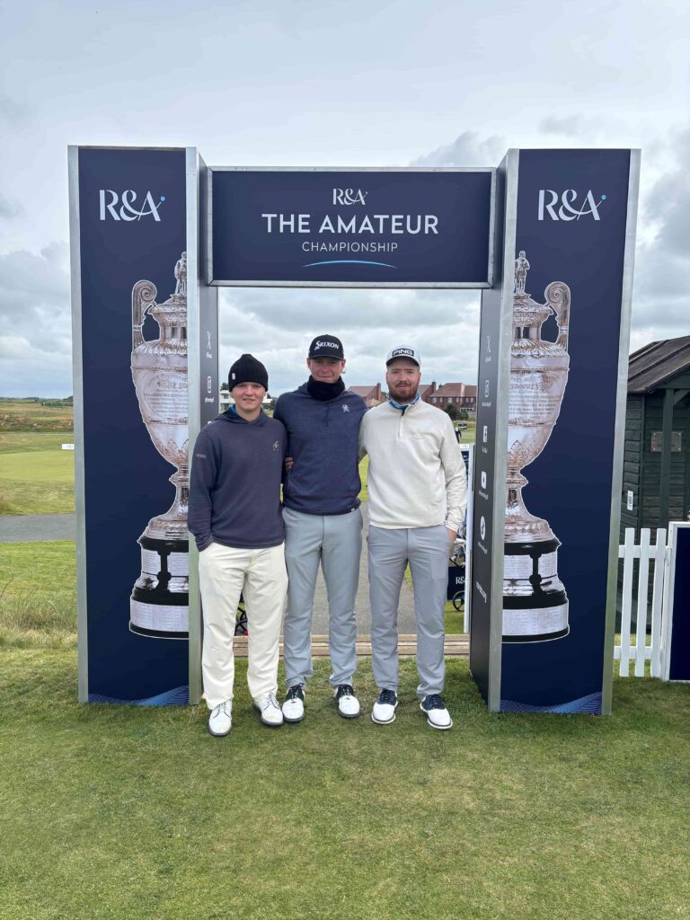 Three male golfers stand together in front of a large blue archway sign for the R&A The Amateur Championship at a golf course.