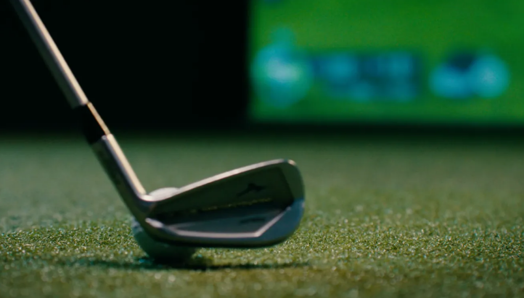 Close-up of a golf putter head on a green putting surface with a blurred practice screen in the background.