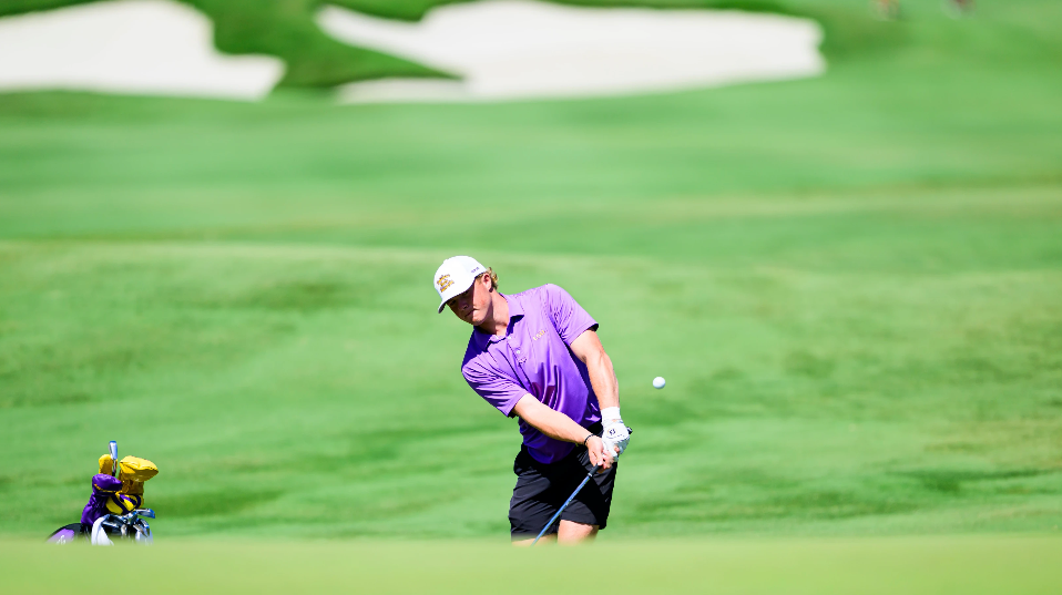 Male golfer in a white cap and purple shirt mid-swing on a lush green golf course, with a bag of clubs nearby.