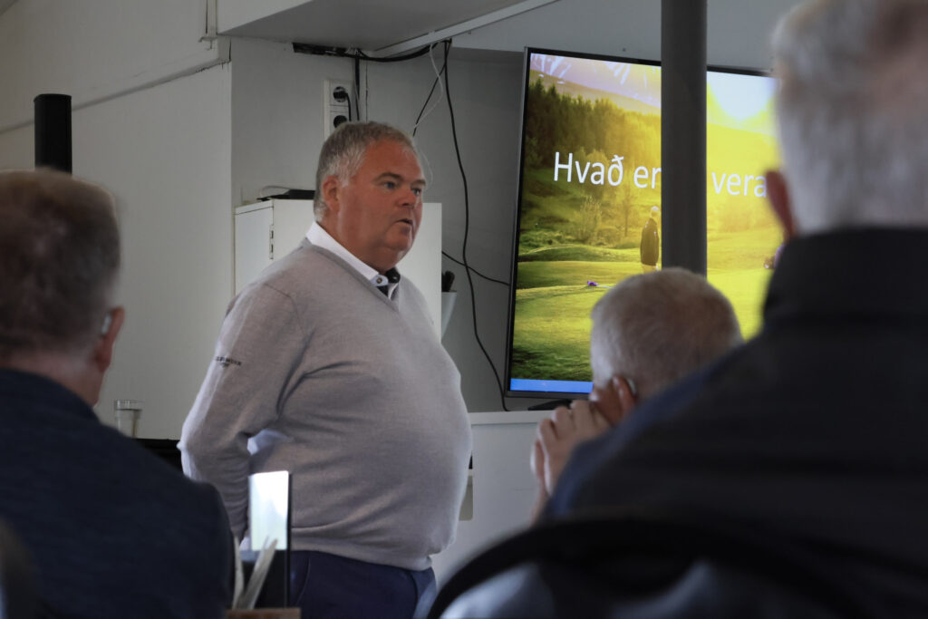 Man in a light gray sweater speaks to an audience in a classroom/meeting room; a large screen shows a nature landscape with text in Danish (Hvad er...).