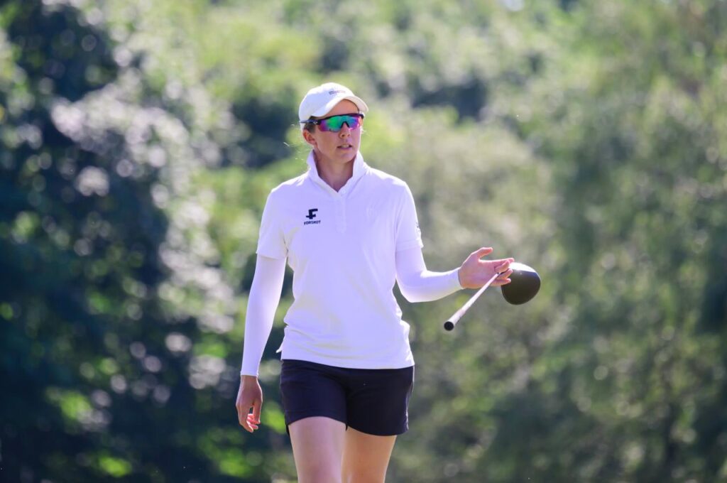 Female golfer in a white polo and cap practices with a tethered ball outdoors in bright sunlight.