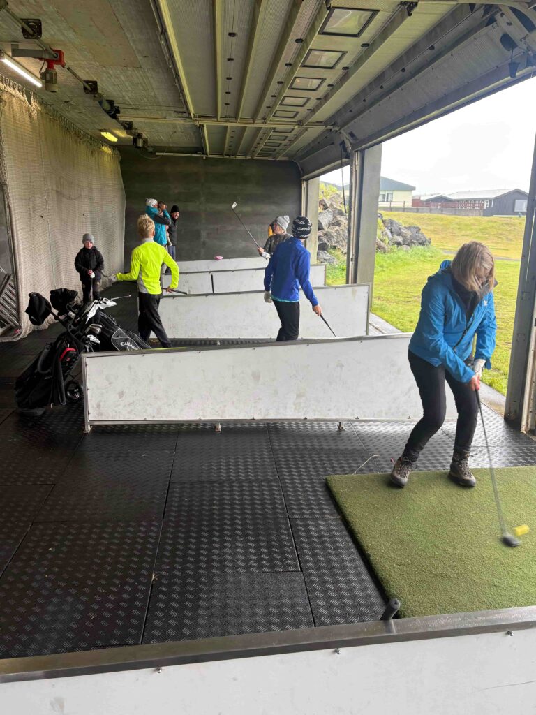 Group of people practicing golf indoors on matting; several players swing clubs near a dark back wall, with a coach or teammate observing in bright jackets.