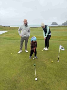 A man smiles beside a woman putting on a seaside golf green with a child watching nearby.
