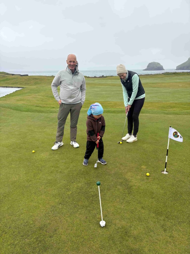 A man smiles beside a woman putting on a seaside golf green with a child watching nearby.