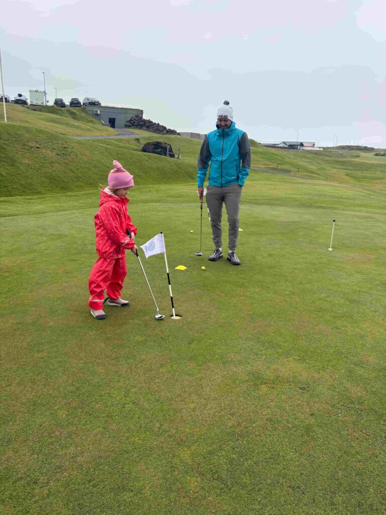 Man in a blue jacket and gray pants watches a child in a pink snowsuit practicing putts on a golf green.