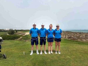 Four young golfers standing in a row on a sunny coastal golf course, wearing matching blue polo shirts and white caps.