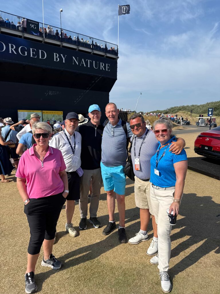 Group of six adults posing together in front of a large dark ship with 'FORGED BY NATURE' on its side, at an outdoor event on a sunny day.