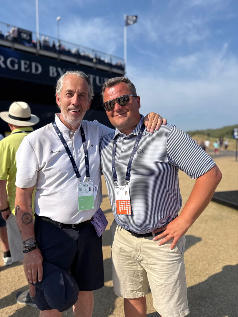 Two men posing for a photo at an outdoor event, wearing lanyards with badges and smiling, with a large ship and blue sky in the background.