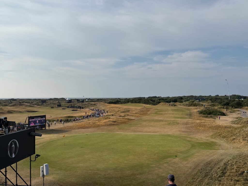 Crowd walking along a fairway toward a stage and large screens at a coastal golf tournament under a blue sky.
