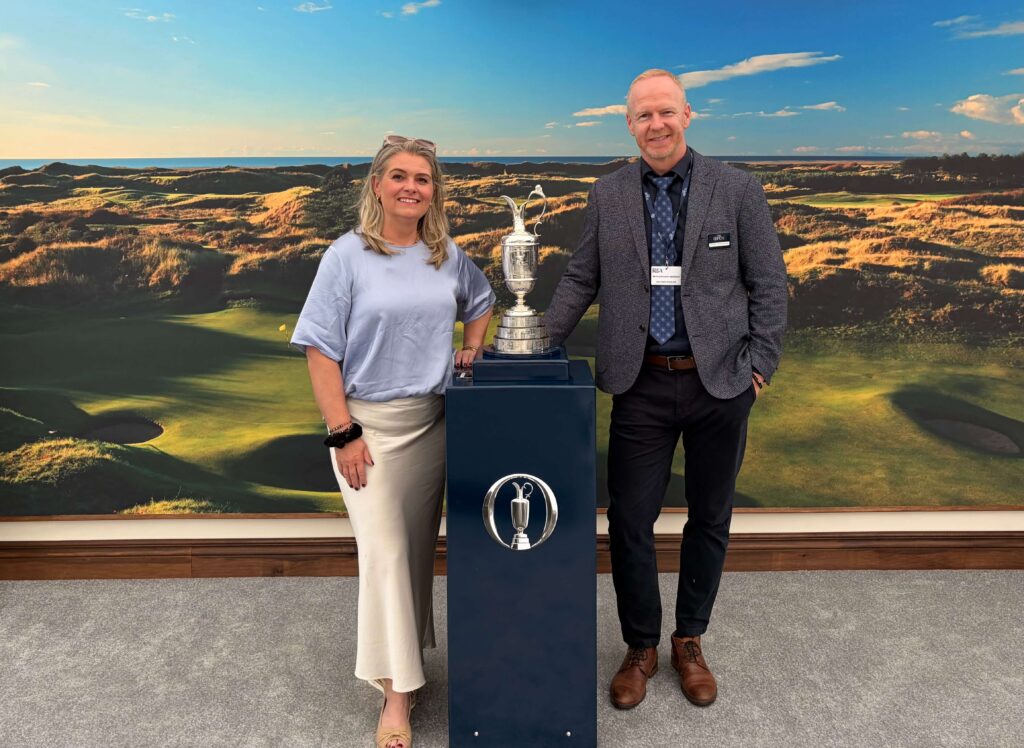Two people stand beside a tall silver golf trophy, posing in front of a golf course backdrop with blue sky.