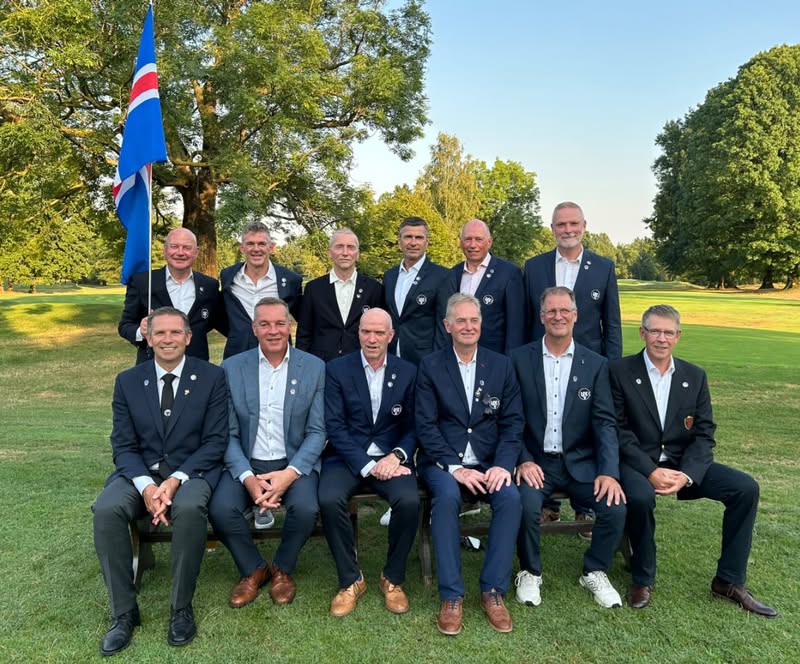 Twelve men in dark suits posing for a formal group photo on a sunny, grassy park; one man holds a blue flag with red and white accents.