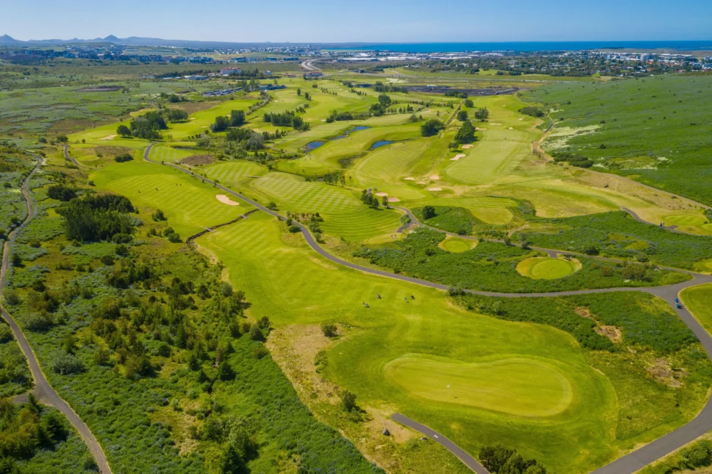 Aerial view of a golf course with greens, fairways, bunkers, and winding cart paths across rolling hills toward the coastline.